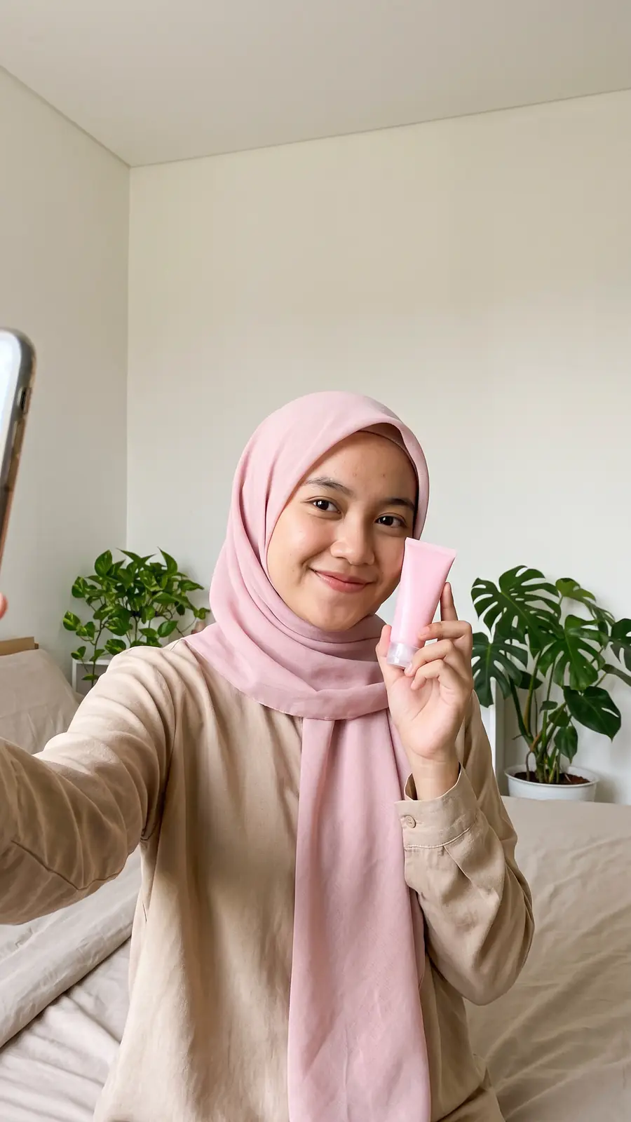 Kreator fiktif perempuan berhijab merah muda berswafoto di kamar terang sambil memegang tube skincare merah muda tanpa label.